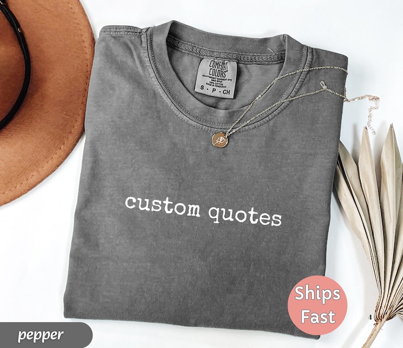 K&ouml;nnte beinhalten: Ein graues T-Shirt mit den wei&szlig;en Worten "custom quotes". Eine goldene Halskette mit Anh&auml;nger liegt auf dem Shirt. Das Shirt ist gefaltet und hat ein Etikett mit der Aufschrift "Comfort Colors". Ein brauner Hut ist im Hintergrund.