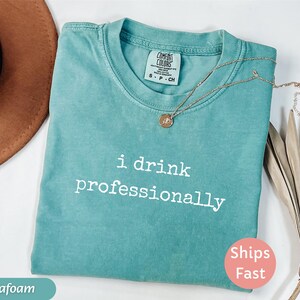 May include: Seafoam green t-shirt with the text "i drink professionally" in white. A gold necklace with a pendant rests on the shirt. The shirt has a crew neck and short sleeves. A "Ships Fast" sticker is visible.