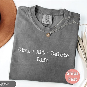 May include: A gray t-shirt with the text "Ctrl + Alt + Delete Life" in white. The shirt has a crew neck and short sleeves. A gold necklace with a pendant is on the shirt. The shirt is folded and has a tag that says "Comfort Colors".