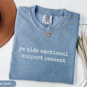 May include: Blue jean colored t-shirt with the text "ye olde emotional support peasant" in white. A gold necklace with a pendant is draped across the shirt. The shirt is folded and has a Comfort Colors tag.