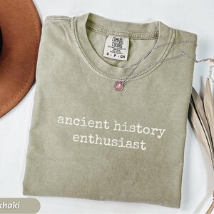 Pu&ograve; includere: T-shirt color cachi con la scritta bianca "ancient history enthusiast". Una collana d'argento con un ciondolo rosa &egrave; appoggiata sulla maglietta. La maglietta ha uno scollo tondo e un'etichetta con la scritta "Comfort Colors".