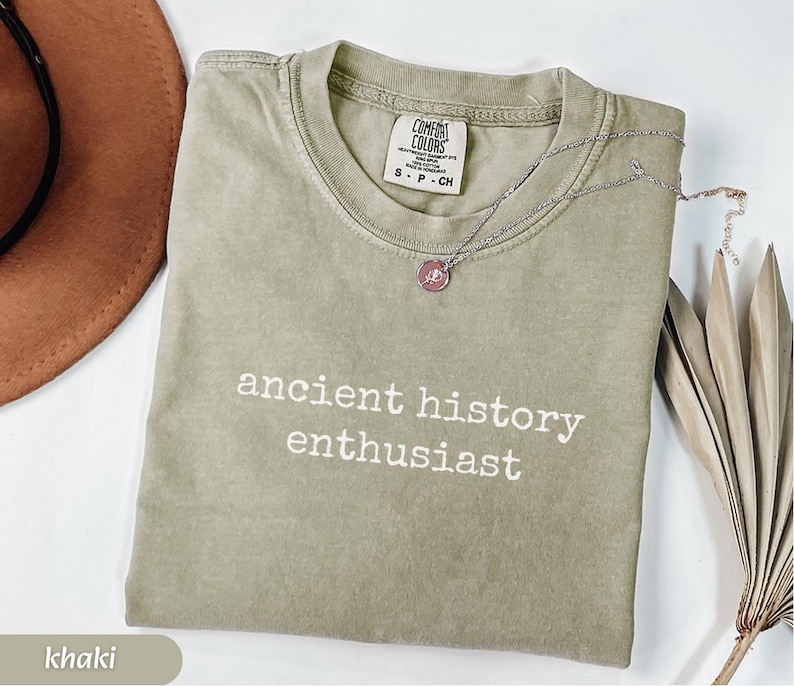 Pu&ograve; includere: T-shirt color cachi con la scritta bianca "ancient history enthusiast". Una collana d'argento con un ciondolo rosa &egrave; appoggiata sulla maglietta. La maglietta ha uno scollo tondo e un'etichetta con la scritta "Comfort Colors".