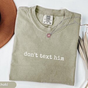 May include: Khaki-colored t-shirt with the phrase "don't text him" in white lettering. The shirt has a crew neck and short sleeves. A silver necklace with a pendant is draped on the shirt. A brown hat and dried plant are in the background.