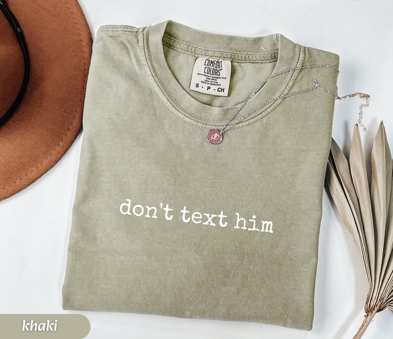 May include: Khaki-colored t-shirt with the phrase "don't text him" in white lettering. The shirt has a crew neck and short sleeves. A silver necklace with a pendant is draped on the shirt. A brown hat and dried plant are in the background.
