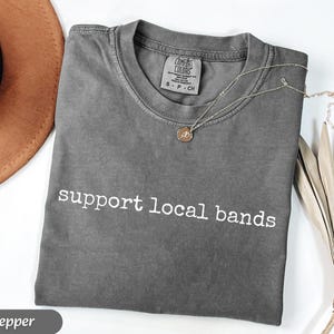 May include: A gray Comfort Colors t-shirt with the text "support local bands" in white. A silver necklace with a small pendant rests on the shirt. A brown hat and dried plant are in the background.