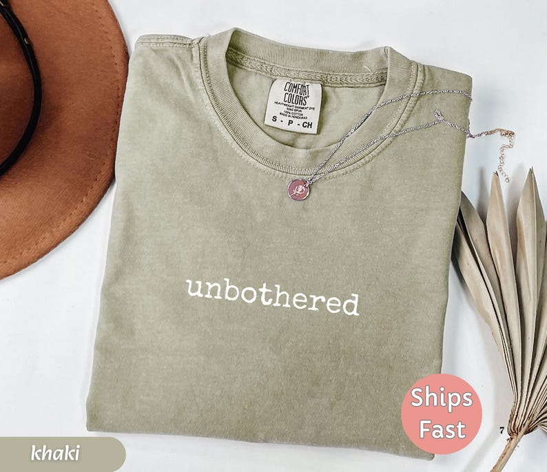 May include: Khaki-colored t-shirt with the word "unbothered" printed in white. The shirt has a crew neck and short sleeves. A silver necklace with a pendant is draped across the shirt. A brown hat is visible in the upper left corner.
