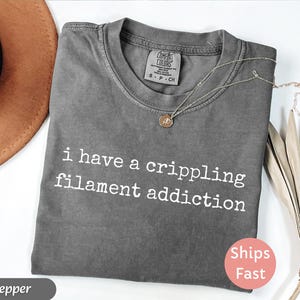 May include: A gray Comfort Colors t-shirt with the text "i have a crippling filament addiction" in white. A gold necklace with a pendant rests on the shirt. A brown hat and dried leaves are in the background.