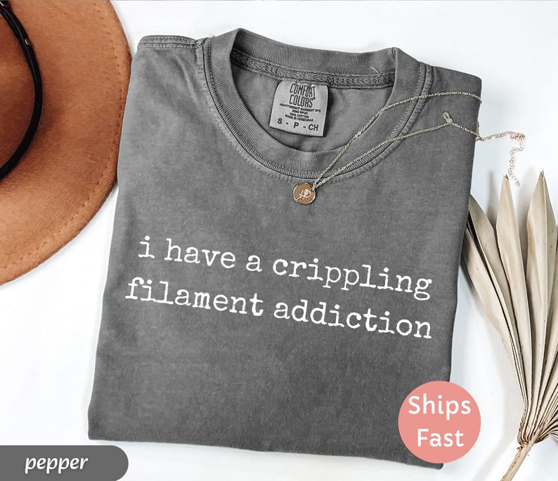 May include: A gray Comfort Colors t-shirt with the text "i have a crippling filament addiction" in white. A gold necklace with a pendant rests on the shirt. A brown hat and dried leaves are in the background.