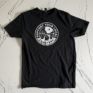 May include: A black short-sleeve t-shirt featuring a white circular graphic on the chest. The design depicts a cemetery scene with tombstones, a building, and a crescent moon, surrounded by the text SUPPORT YOUR LOCAL CEMETERY in a bold font.