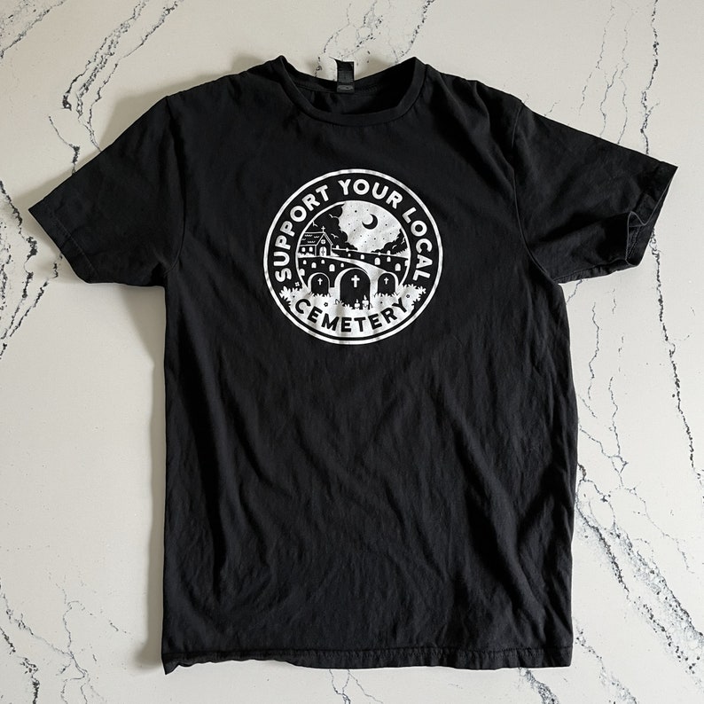 May include: A black short-sleeve t-shirt featuring a white circular graphic on the chest. The design depicts a cemetery scene with tombstones, a building, and a crescent moon, surrounded by the text SUPPORT YOUR LOCAL CEMETERY in a bold font.