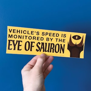 May include: Yellow bumper sticker with black text that reads "VEHICLE'S SPEED IS MONITORED BY THE EYE OF SAURON." The sticker features a graphic of the Eye of Sauron. The background is a solid blue.