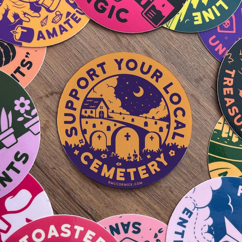 May include: A round sticker with a purple background and yellow accents. The sticker features a silhouette of a church with a crescent moon and stars in the sky. The text "Support Your Local Cemetery" is written in a circle around the image. The website "BMCCORMICK.COM" is printed at the bottom of the sticker.