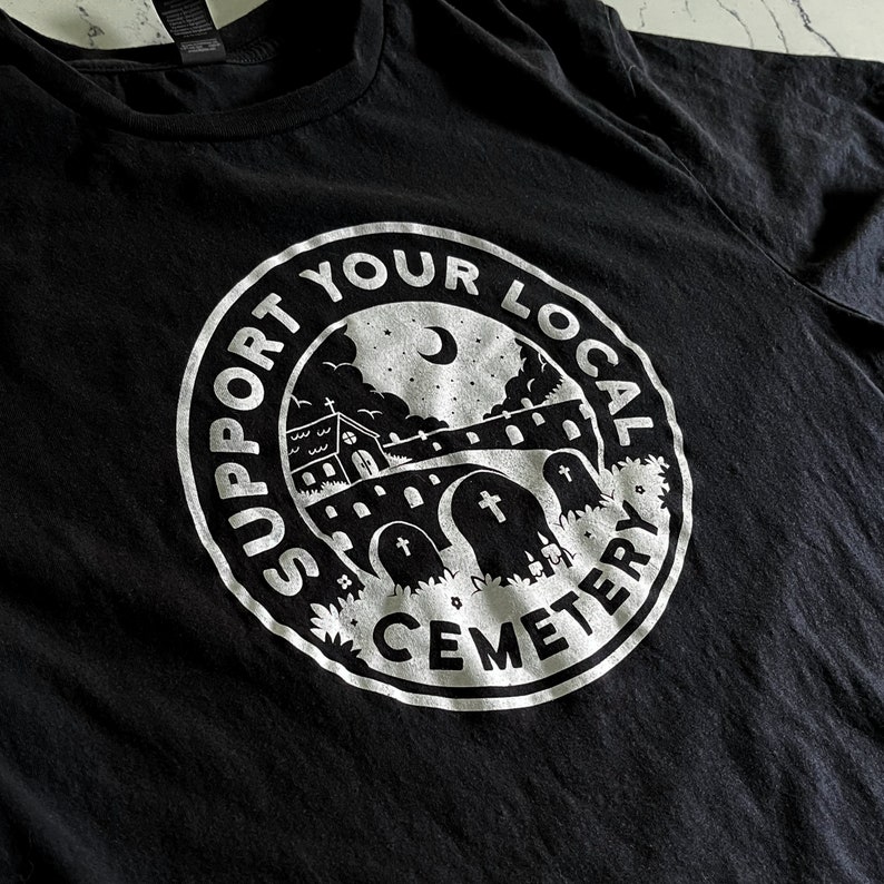 May include: A black t-shirt featuring a circular white graphic print. The design depicts a cemetery scene with tombstones, a small building, a crescent moon, and stars. The text "SUPPORT YOUR LOCAL CEMETERY" is printed in a circular border.