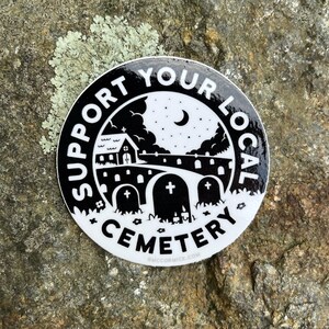 May include: Black and white sticker with the text "Support Your Local Cemetery" in a circle around a black and white illustration of a church, a graveyard, and a crescent moon.