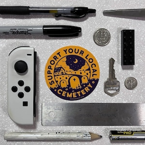 May include: A round yellow sticker with a black outline and the text "Support Your Local Cemetery" in a black circle. The sticker features a black silhouette of a cemetery with a church, a crescent moon, and stars. The website address "EMCCORMICE.COM" is printed in black at the bottom of the sticker.