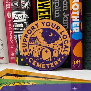 May include: A round sticker with an orange background and purple text. The text reads "Support Your Local Cemetery" with a small church, a crescent moon, and three tombstones in the center. The bottom of the sticker reads "BMCCORMICK.COM".