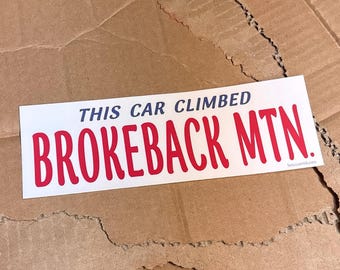 May include: A rectangular white bumper sticker with dark blue text that reads THIS CAR CLIMBED and large red text that reads BROKEBACK MTN. The sticker also features small black text in the bottom right corner that reads breccomick.com.