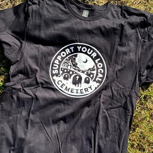 May include: A black short-sleeve t-shirt featuring a circular white graphic on the chest. The design depicts a cemetery scene with a church, gravestones, and a crescent moon, surrounded by the text SUPPORT YOUR LOCAL CEMETERY in a bold font.