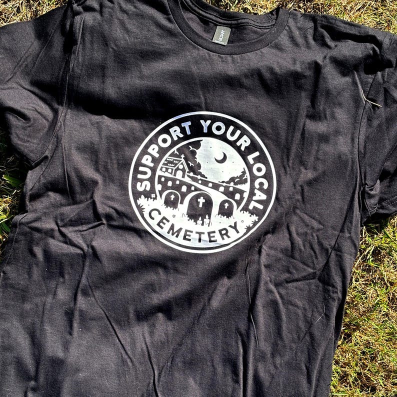 May include: A black short-sleeve t-shirt featuring a circular white graphic on the chest. The design depicts a cemetery scene with a church, gravestones, and a crescent moon, surrounded by the text SUPPORT YOUR LOCAL CEMETERY in a bold font.
