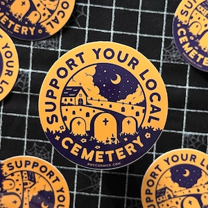 May include: A round sticker with a yellow background and purple text. The text reads "Support Your Local Cemetery". The sticker features a cartoon illustration of a church, a graveyard, and a crescent moon in the night sky.