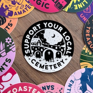 May include: Black and white sticker with the text "Support Your Local Cemetery" in a circle. The sticker features a small church, a crescent moon, and a graveyard with three tombstones.