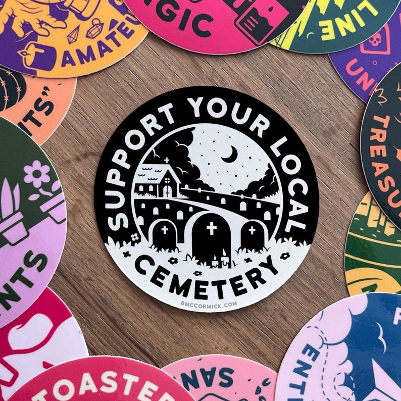 May include: Black and white sticker with the text "Support Your Local Cemetery" in a circle. The sticker features a small church, a crescent moon, and a graveyard with three tombstones.