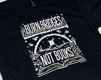 May include: A black short-sleeve t-shirt featuring a white graphic print. The design depicts a burning stone bridge above an open book, with the text "BURN BRIDGES NOT BOOKS" printed in a bold, stylized font. The illustration includes stars and flames.