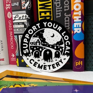 May include: Black and white sticker with the text "Support Your Local Cemetery" in a circle. The sticker features a small church, a crescent moon, and a graveyard with tombstones.