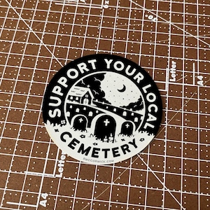 May include: Black and white sticker with the text "Support Your Local Cemetery" in a circle. The sticker features a small church, a bridge, and tombstones under a starry night sky.