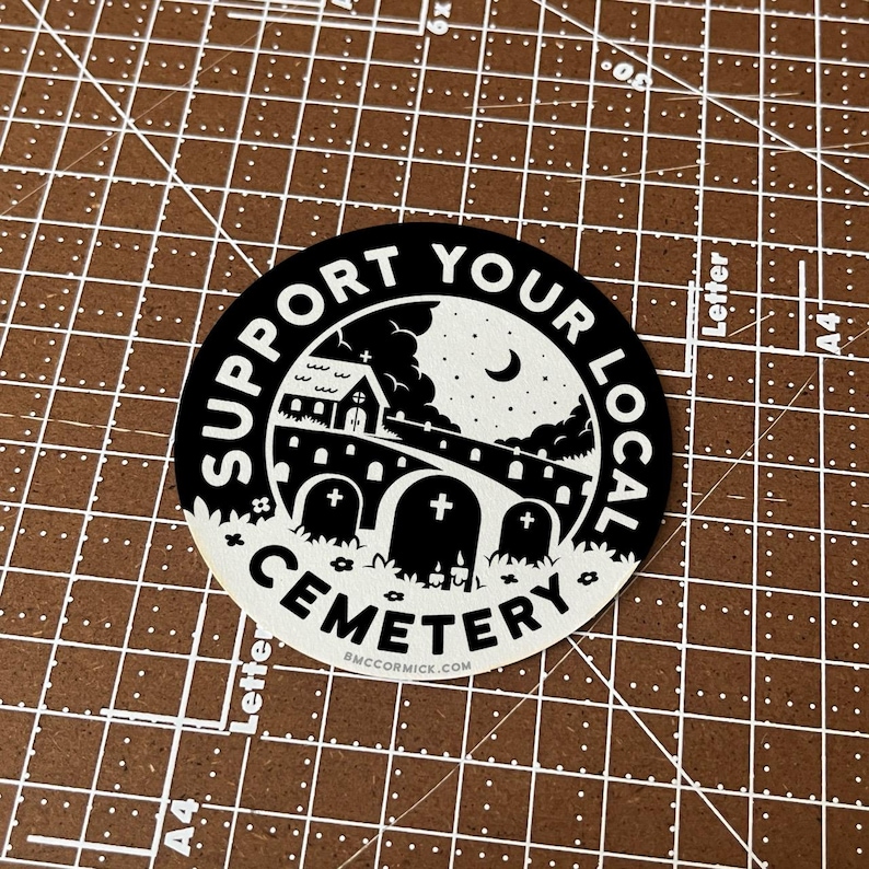 May include: Black and white sticker with the text "Support Your Local Cemetery" in a circle. The sticker features a small church, a bridge, and tombstones under a starry night sky.