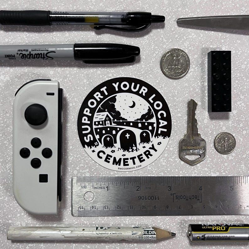 May include: A black and white sticker with the text "Support Your Local Cemetery" and a silhouette of a graveyard with a church and a crescent moon. The sticker is on a white background.