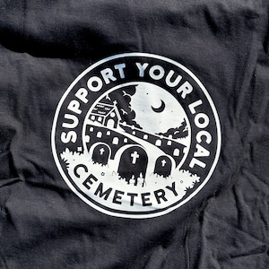 May include: A black t-shirt featuring a white circular graphic print. The design depicts a cemetery scene with tombstones, a building, and a crescent moon under the text "SUPPORT YOUR LOCAL CEMETERY" arranged in a circular border around the illustration.