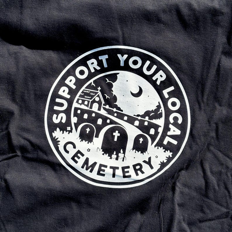 May include: A black t-shirt featuring a white circular graphic print. The design depicts a cemetery scene with tombstones, a building, and a crescent moon under the text "SUPPORT YOUR LOCAL CEMETERY" arranged in a circular border around the illustration.