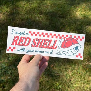 May include: A rectangular white bumper sticker featuring the text I've got a RED SHELL with your name on it in blue and red lettering. The design includes a red and white checkered border and a graphic illustration of a red turtle shell.