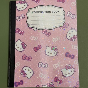 Puede incluir: Cuaderno de composición rosa con un patrón de caras y lazos de Hello Kitty. La portada tiene las palabras "COMPOSITION BOOK" y líneas para un nombre. El cuaderno tiene un lomo negro y un acabado brillante.