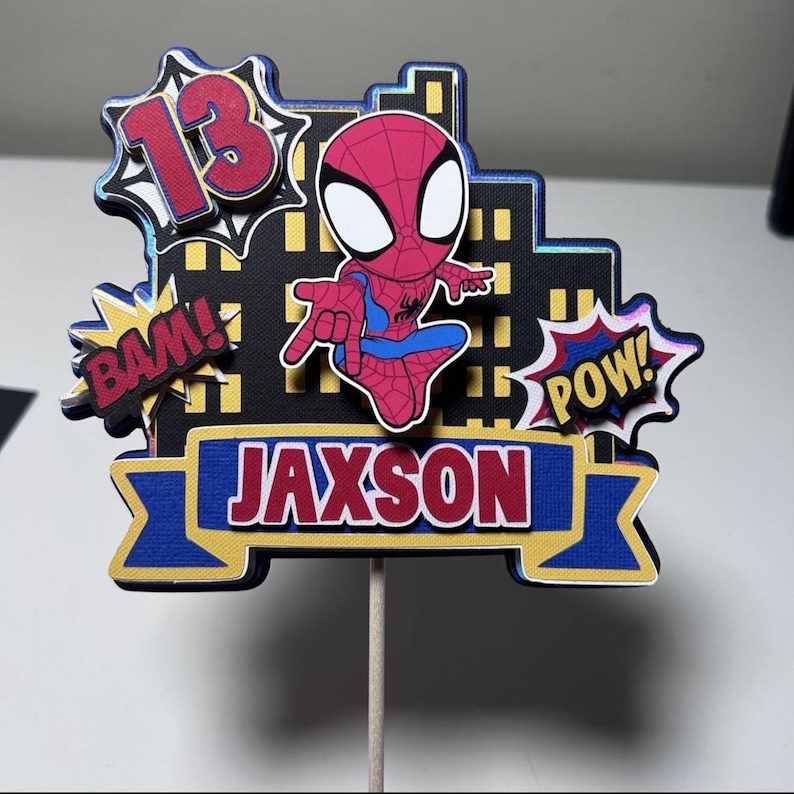 May include: A colorful cake topper featuring a cartoon Spider-Man character. The design includes the number "13" in a comic book style, the name "Jaxson", and action words "BAM!" and "POW!" on a cityscape background. The topper is mounted on a wooden stick.