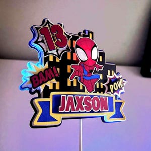 May include: A colorful cake topper featuring Spider-Man, the number 13, and the name "Jaxson." The design includes comic book style speech bubbles with "BAM!" and "POW!" The topper is primarily red, blue, and black.