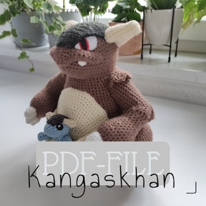 Könnte beinhalten: Gehäkeltes Amigurumi-Spielzeug von Kangama, einem Pokémon-Charakter, das ein kleineres gehäkeltes Spielzeug eines Baby-Kangama hält. Das größere Spielzeug ist braun und beige mit einem schwarzen Kopf und roten Augen. Das kleinere Spielzeug ist blau mit schwarzen Augen. Der Text "PDF-FILE Kangaskhan" ist auf weißem Hintergrund gedruckt.