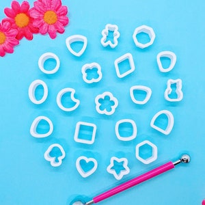 May include: A collection of twenty-one white plastic geometric and organic shape cutters arranged in a circular pattern, including a bear, star, heart, mushroom, and flower, accompanied by a pink metal ball stylus tool for crafting.