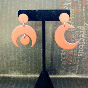 May include: A pair of matte peach-colored earrings featuring a crescent moon shape. One earring consists of a small circular stud attached to a larger crescent moon with a small circle suspended in the center. The other is a simple crescent moon.