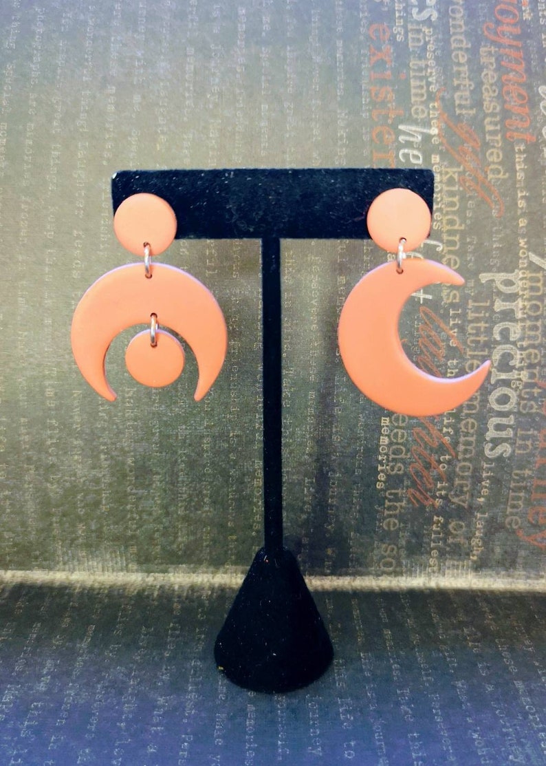 May include: A pair of matte peach-colored earrings featuring a crescent moon shape. One earring consists of a small circular stud attached to a larger crescent moon with a small circle suspended in the center. The other is a simple crescent moon.