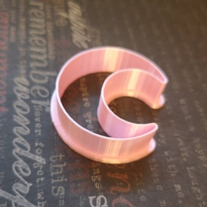 May include: A light pink plastic cookie cutter shaped like a crescent moon. The tool features a wide base rim and a raised, curved cutting edge that forms a distinct moon silhouette, constructed with visible vertical layers from the 3D printing process.