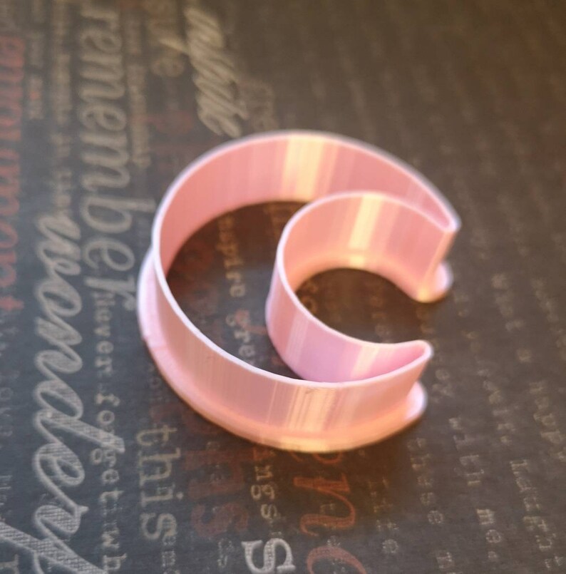 May include: A light pink plastic cookie cutter shaped like a crescent moon. The tool features a wide base rim and a raised, curved cutting edge that forms a distinct moon silhouette, constructed with visible vertical layers from the 3D printing process.