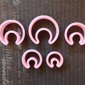 May include: A set of five light pink plastic cookie cutters, each featuring a crescent moon shape. The cutters are arranged in a group with three larger pieces positioned across the top and two smaller pieces placed below them on a dark background.