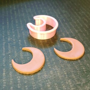 May include: A light pink crescent moon-shaped cookie cutter made of plastic, accompanied by two flat, light pink crescent moon-shaped cutouts. The items are arranged on a dark surface with faint, repeating text patterns visible in the background.