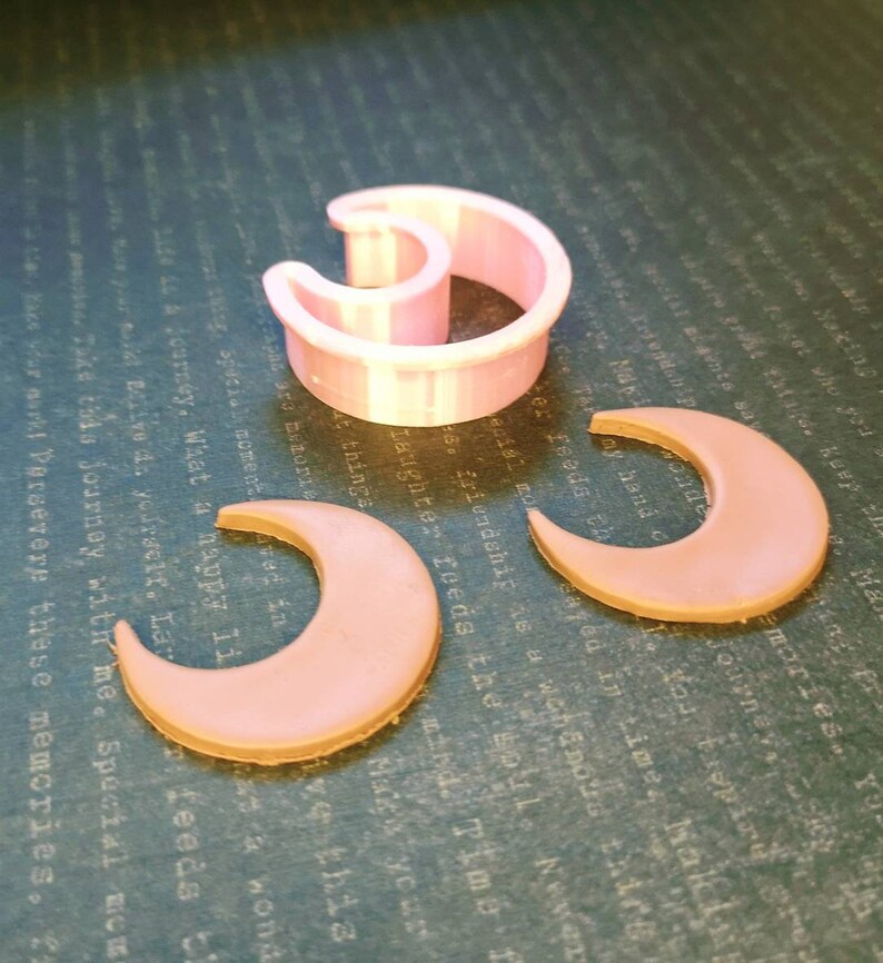 May include: A light pink crescent moon-shaped cookie cutter made of plastic, accompanied by two flat, light pink crescent moon-shaped cutouts. The items are arranged on a dark surface with faint, repeating text patterns visible in the background.