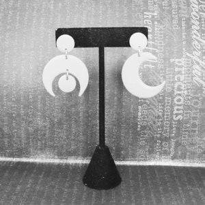 May include: A pair of white earrings displayed on a black T-shaped stand. The left earring features a circular stud connected to a crescent moon shape with a small circle suspended inside. The right earring consists of a circular stud and a crescent moon.