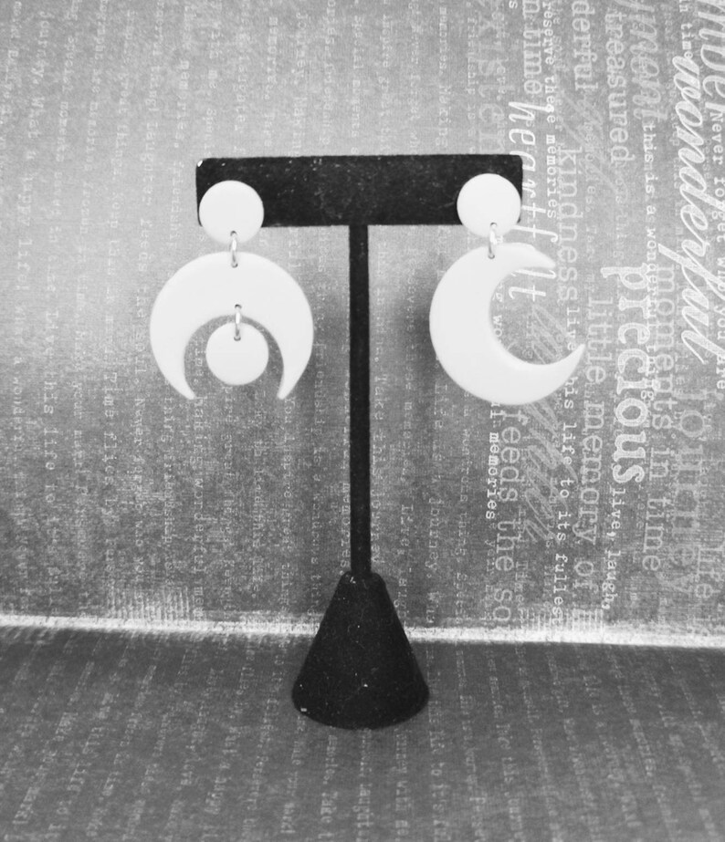 May include: A pair of white earrings displayed on a black T-shaped stand. The left earring features a circular stud connected to a crescent moon shape with a small circle suspended inside. The right earring consists of a circular stud and a crescent moon.