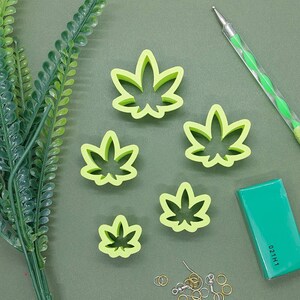 May include: Five light green cannabis leaf-shaped cookie cutters of varying sizes, a green dotting tool, a rectangular green block with the text 021H1, and several small silver and gold metal jump rings and earring hooks for jewellery making.