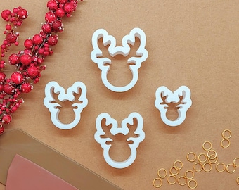 May include: Four white reindeer-shaped cookie cutters of varying sizes arranged in a cluster. The cutters feature a simple outline of a reindeer head with antlers. Small gold-colored metal jump rings are scattered to the right of the cutters.